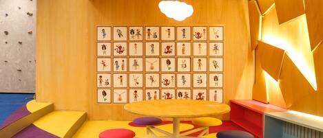 A playroom with a climbing wall, colorful play mats, a round table with stools, and a wall with framed illustrations.
