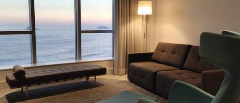 A modern living room with a large window overlooking the sea, a brown sofa, a green armchair, and a small bench.