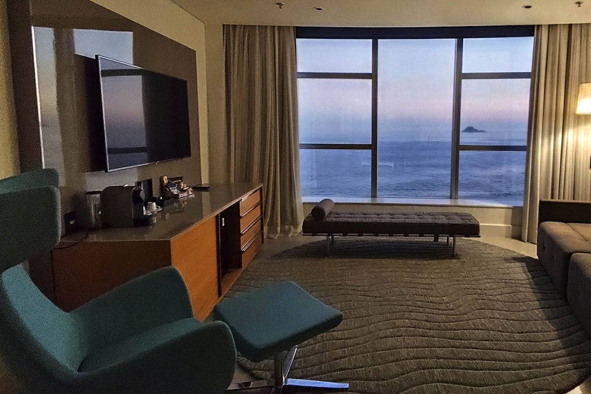 A modern hotel room with a large window offering a view of the sea, a flat-screen TV, a sofa, a chair, and a small table.