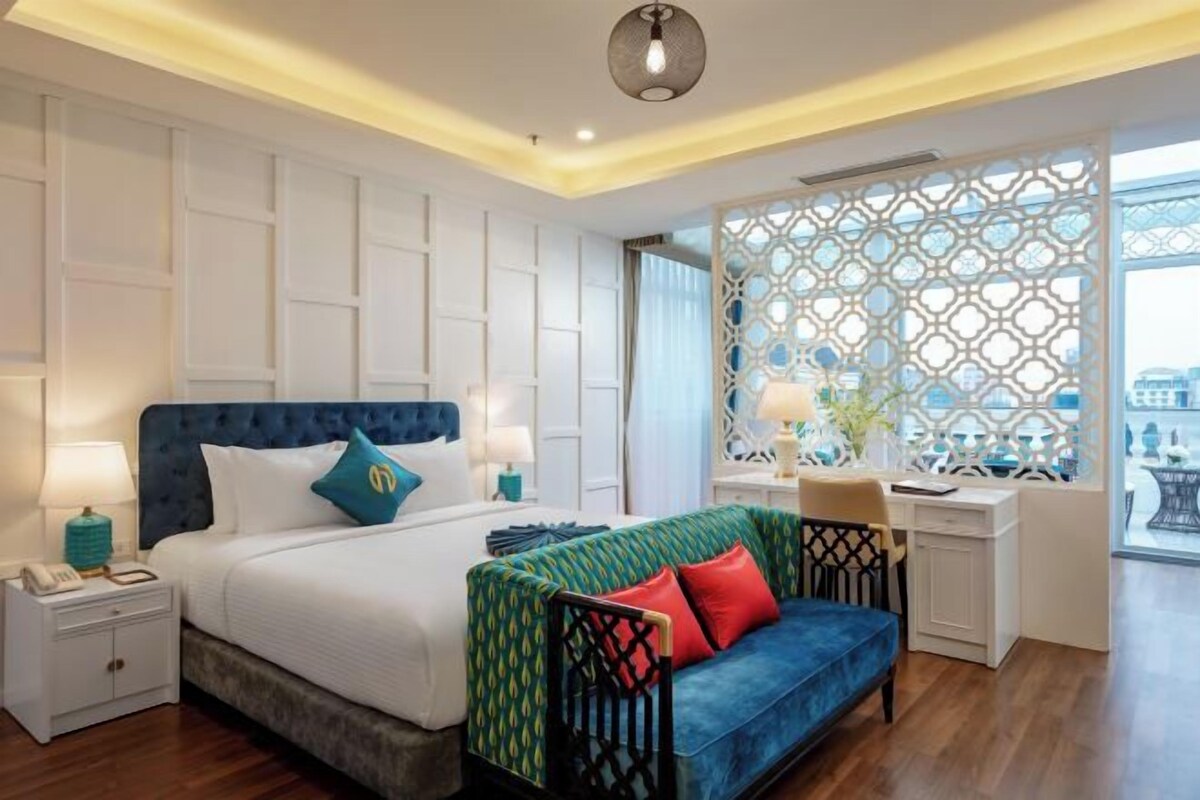 A modern hotel room with a large bed, a desk, and a view through a patterned window.
