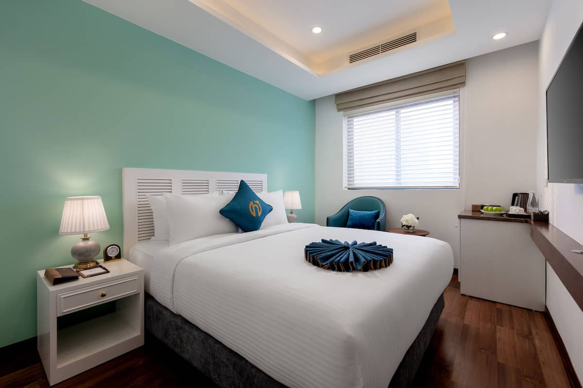 Quarto de hotel moderno com cama, mesa de cabeceira, abajur e uma pequena escrivaninha com cadeira.