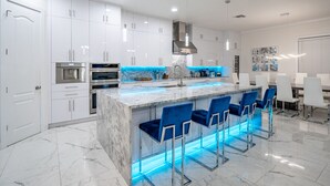 A modern kitchen with white cabinets, a marble countertop, and blue bar stools. - ⭐️S. Tampa Mansion🔥Heated Pool+Spa🎬Movie Theater Chef’s Kitchen 5000sqft (Tampa)