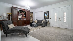 A living room with a dark armchair, a sofa, a coffee table, a bookshelf, and a chandelier. - ⭐️S. Tampa Mansion🔥Heated Pool+Spa🎬Movie Theater Chef’s Kitchen 5000sqft (Tampa)