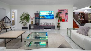 A modern living room with a flat-screen TV, a glass coffee table, a white sofa, and a bookshelf. - ⭐️S. Tampa Mansion🔥Heated Pool+Spa🎬Movie Theater Chef’s Kitchen 5000sqft (Tampa)
