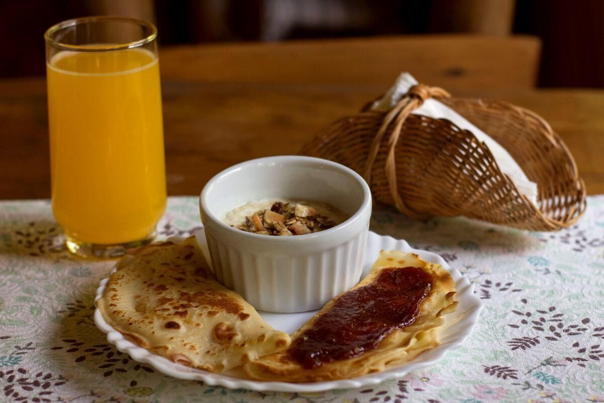 A plate with pancakes and a small bowl of granola, a glass of orange juice, and a basket on a tablecloth.