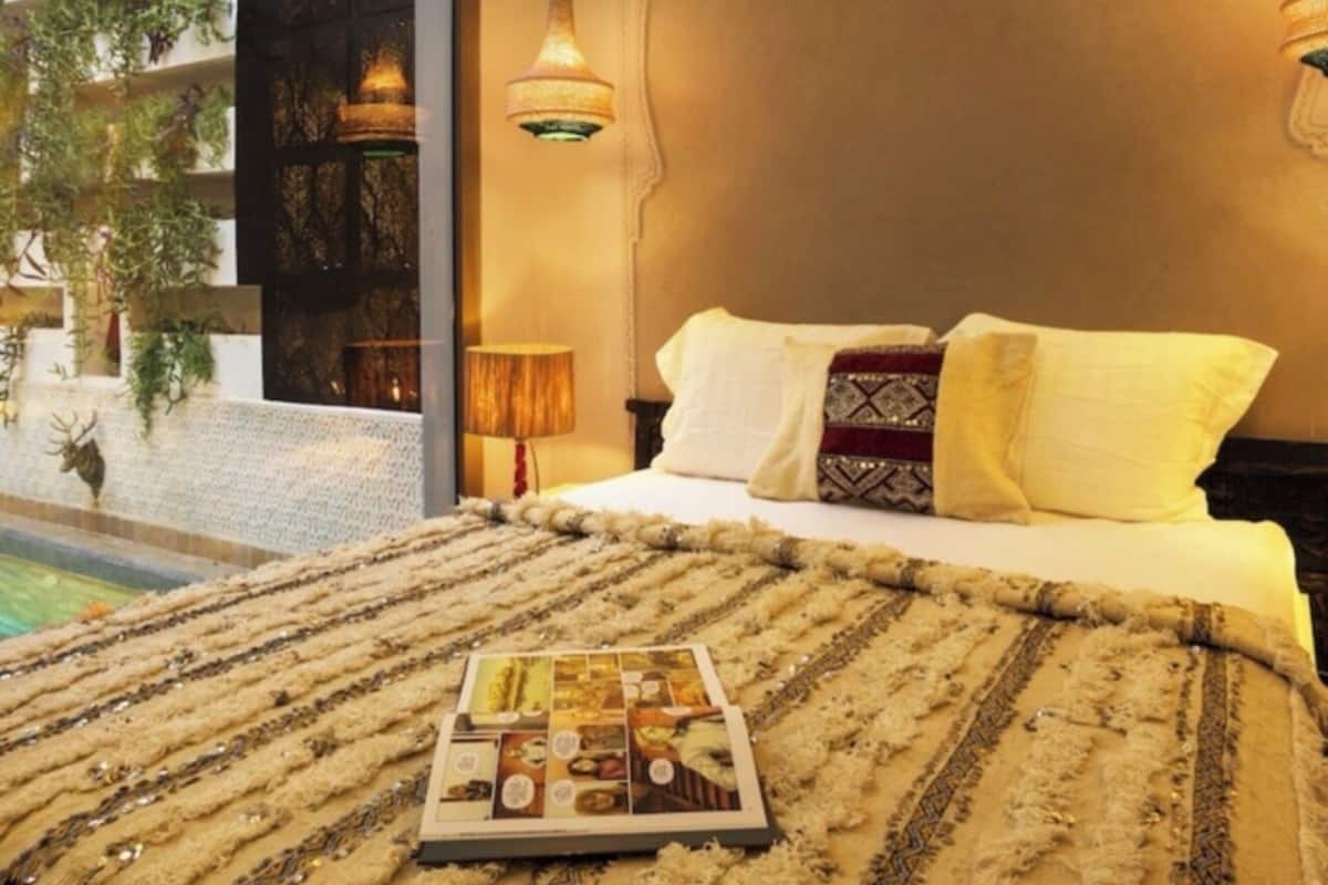 Quarto com cama, uma revista sobre a cama, luminária e vista para uma piscina e vegetação lá fora.