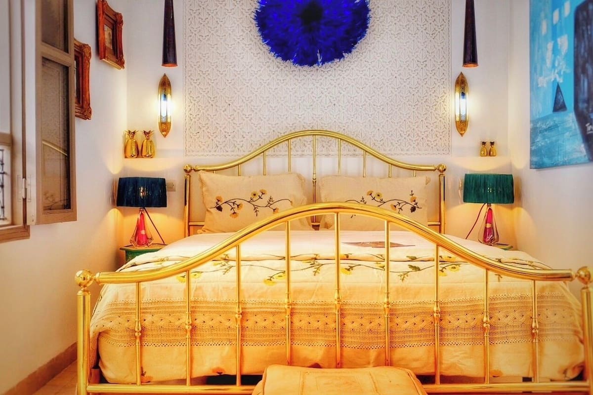 Uma cama com estrutura dourada e roupa de cama florida, arandelas na parede, um ornamento circular azul e quadros emoldurados.