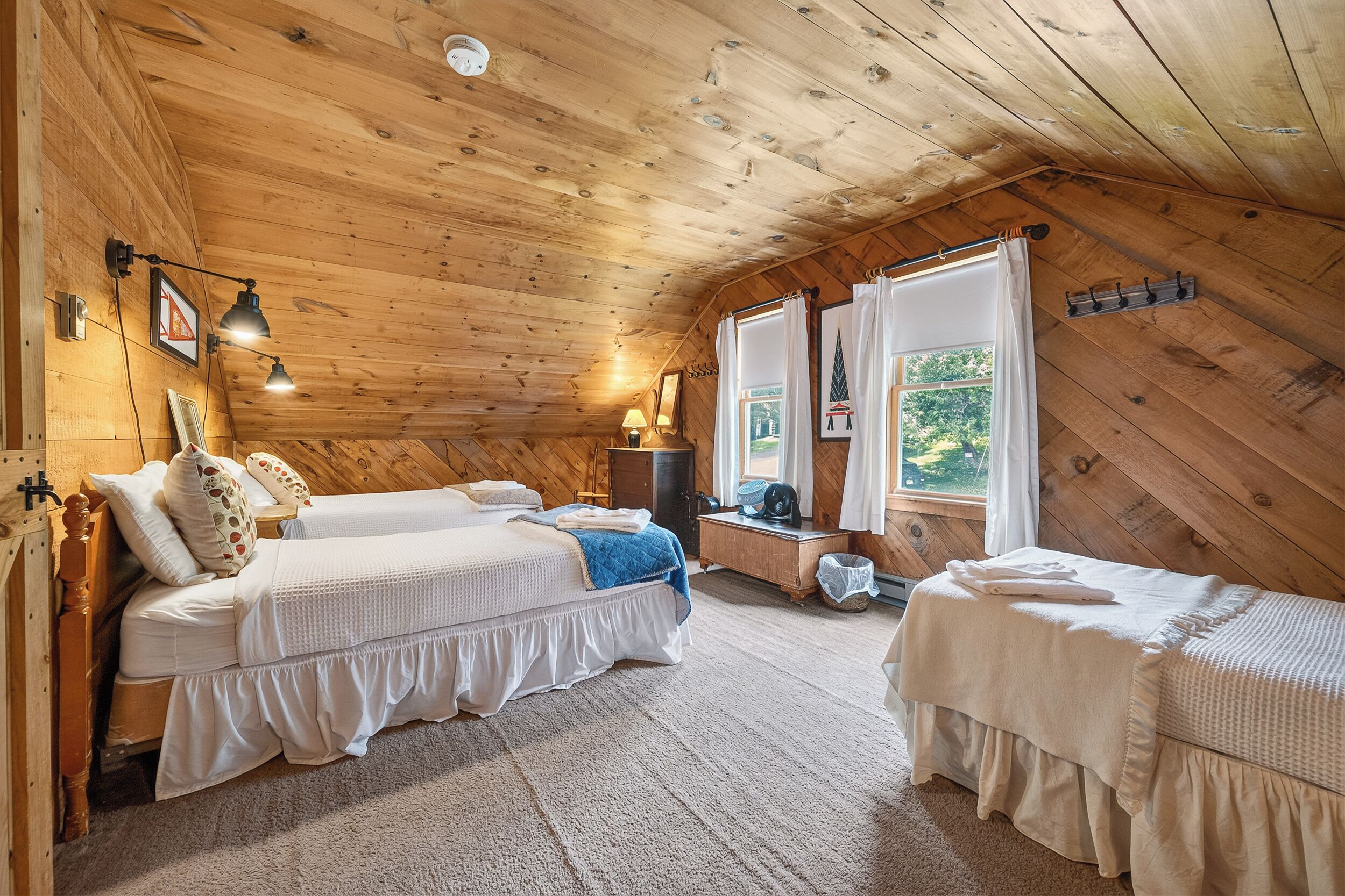 A cozy cabin-style bedroom with two beds, a wooden ceiling, and a view of greenery outside.