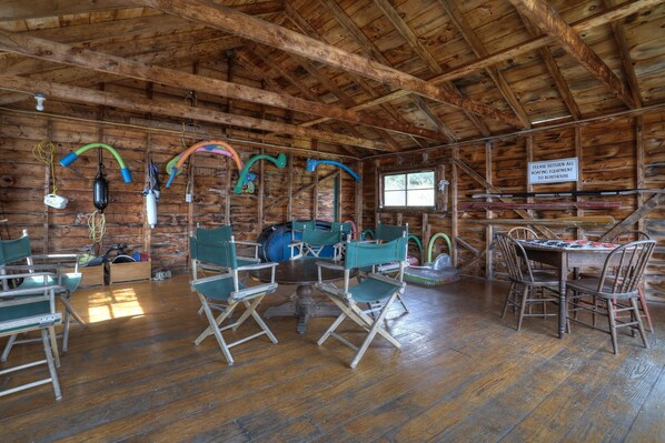 A rustic wooden cabin interior with a dining table, chairs, and various boathouse equipment.
