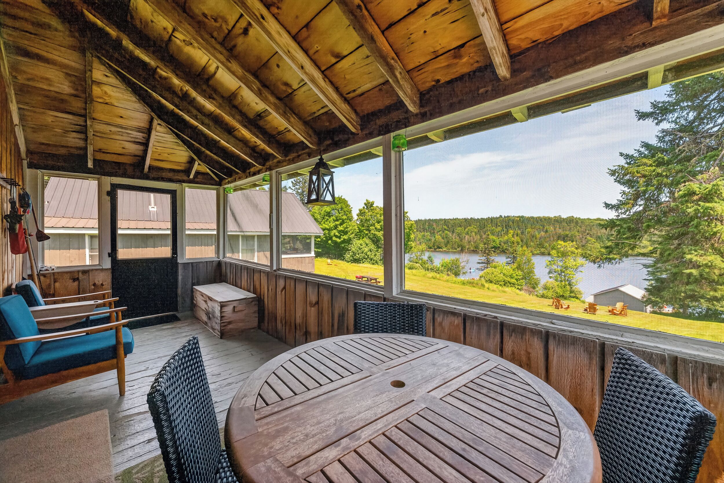 A covered outdoor seating area with a wooden table and chairs, large windows offering a view of a lake and forest.