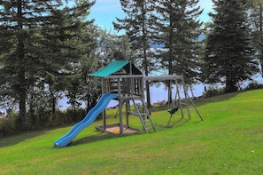 A playground with a slide and swings, surrounded by trees and a grassy area. - Hunter Cove Cabin #4 in the Hunter Cove Cabins Community! (Rangeley)