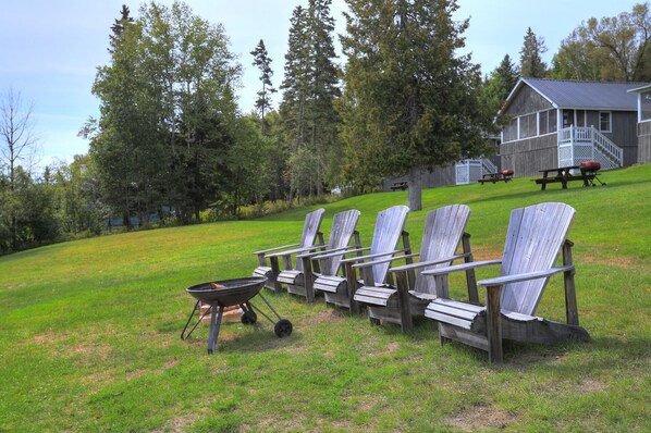 A row of Adirondack chairs on a grassy lawn with a grill and a wooden building in the background.