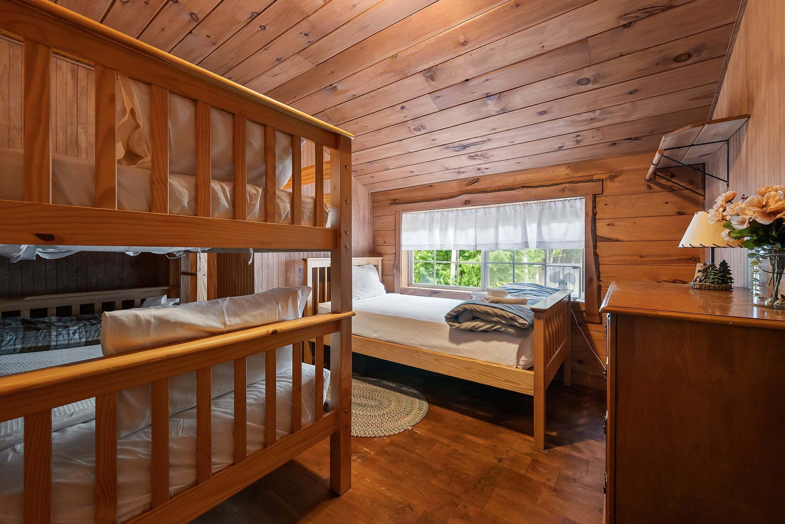 Une chambre dans un chalet en bois, avec un lit superposé, un lit simple, une fenêtre donnant sur un paysage agréable et un bureau avec une lampe et des fleurs.