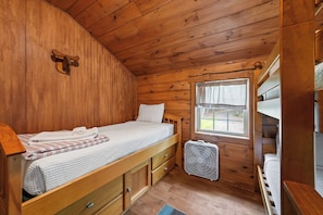 Une petite pièce de cabane en bois comprenant un lit superposé, un lit simple, une fenêtre avec des rideaux et un petit ventilateur.