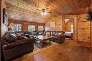 L’intérieur chaleureux d’un chalet avec un plafond en bois, un ventilateur de plafond, une cheminée, des canapés en cuir, une table basse et un lit près d’une fenêtre.