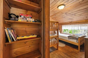 A wooden cabin interior with a bunk bed, a bookshelf with puzzles, and a window with curtains.