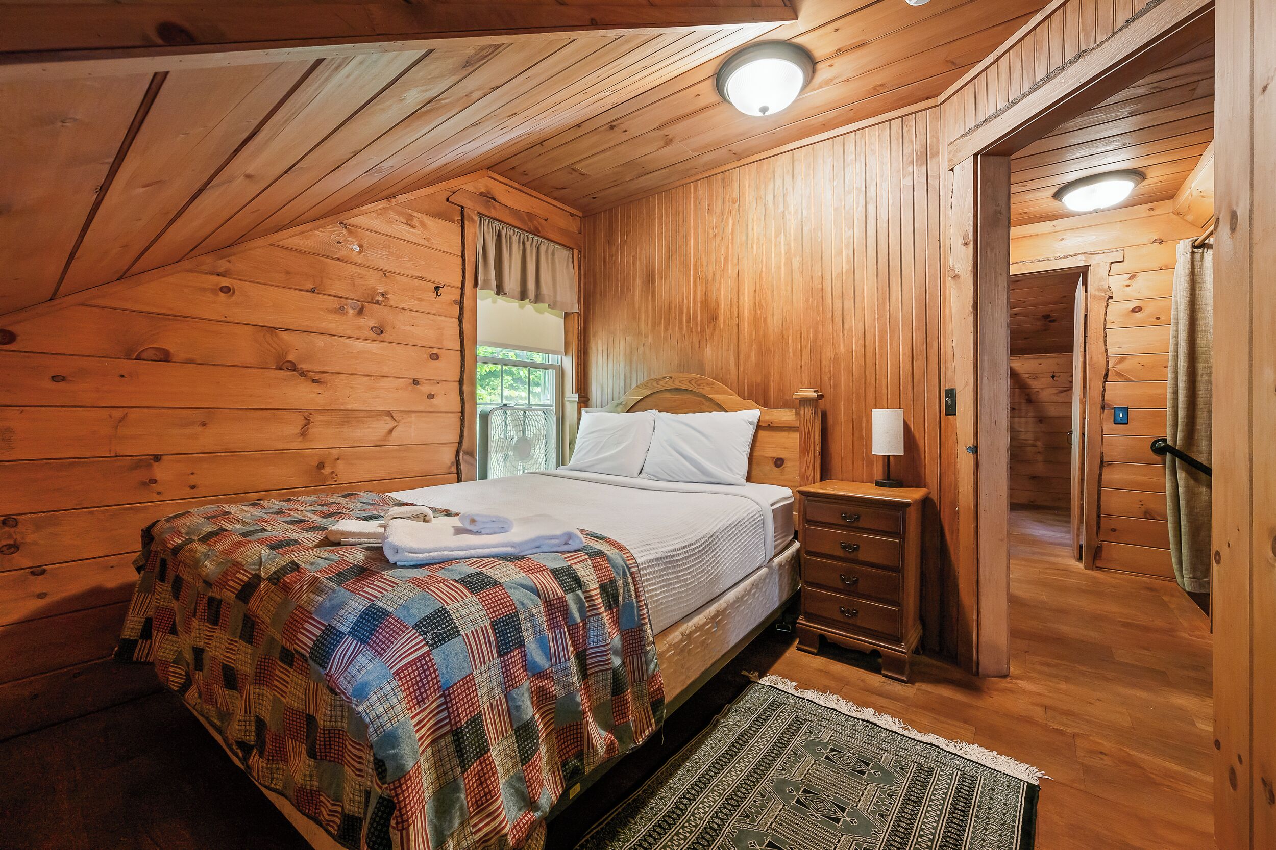 Un dormitori de cabana de fusta amb un llit, una tauleta de nit, una làmpada i una finestra amb cortines.