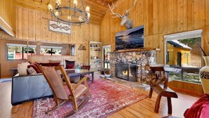 L’intérieur chaleureux d’un chalet avec des murs en bois, une cheminée en pierre, une tête de cerf accrochée au mur et une grande fenêtre donnant sur l’extérieur.