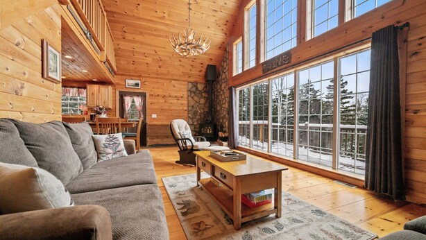 A cozy log cabin interior with a large window, a fireplace, and a dining area. - 🐾Michaud Mountain Chalet - Private cabin with incredible views of Rangeley Lake! Dog Friendly (Rangeley)