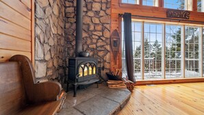 A cozy cabin interior with a stone wall, a wood-burning stove, a bench, and a large window offering a view of trees. - 🐾Michaud Mountain Chalet - Private cabin with incredible views of Rangeley Lake! Dog Friendly (Rangeley)