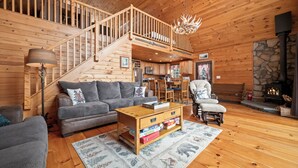 A cozy log cabin interior with a wooden staircase, a fireplace, a dining area, and a living room furnished with a sofa, armchair, and coffee table. - 🐾Michaud Mountain Chalet - Private cabin with incredible views of Rangeley Lake! Dog Friendly (Rangeley)