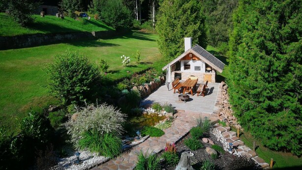A rustic cabin with a stone patio, surrounded by lush greenery and a small pond.