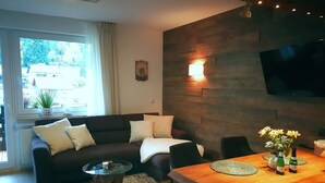 A modern living room with a dark-colored sofa, a wooden dining table, and a flat-screen TV mounted on the wall.