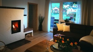 A cozy living room with a fireplace, a sofa, a coffee table with a vase of flowers, and a view of houses through the window.