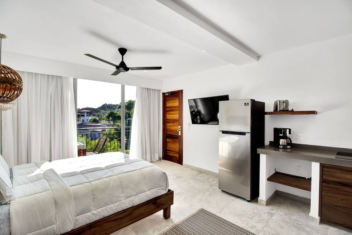 A bedroom with a bed, a ceiling fan, a TV, a refrigerator, and a kitchenette.
