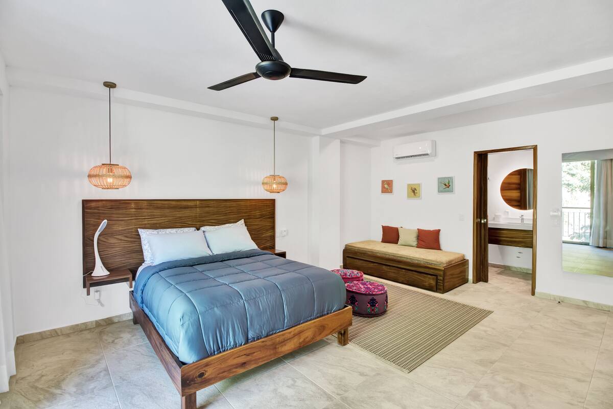 A bedroom with a wooden bed, a ceiling fan, hanging lamps, a bedside table, a bench, and a view of the outdoors.