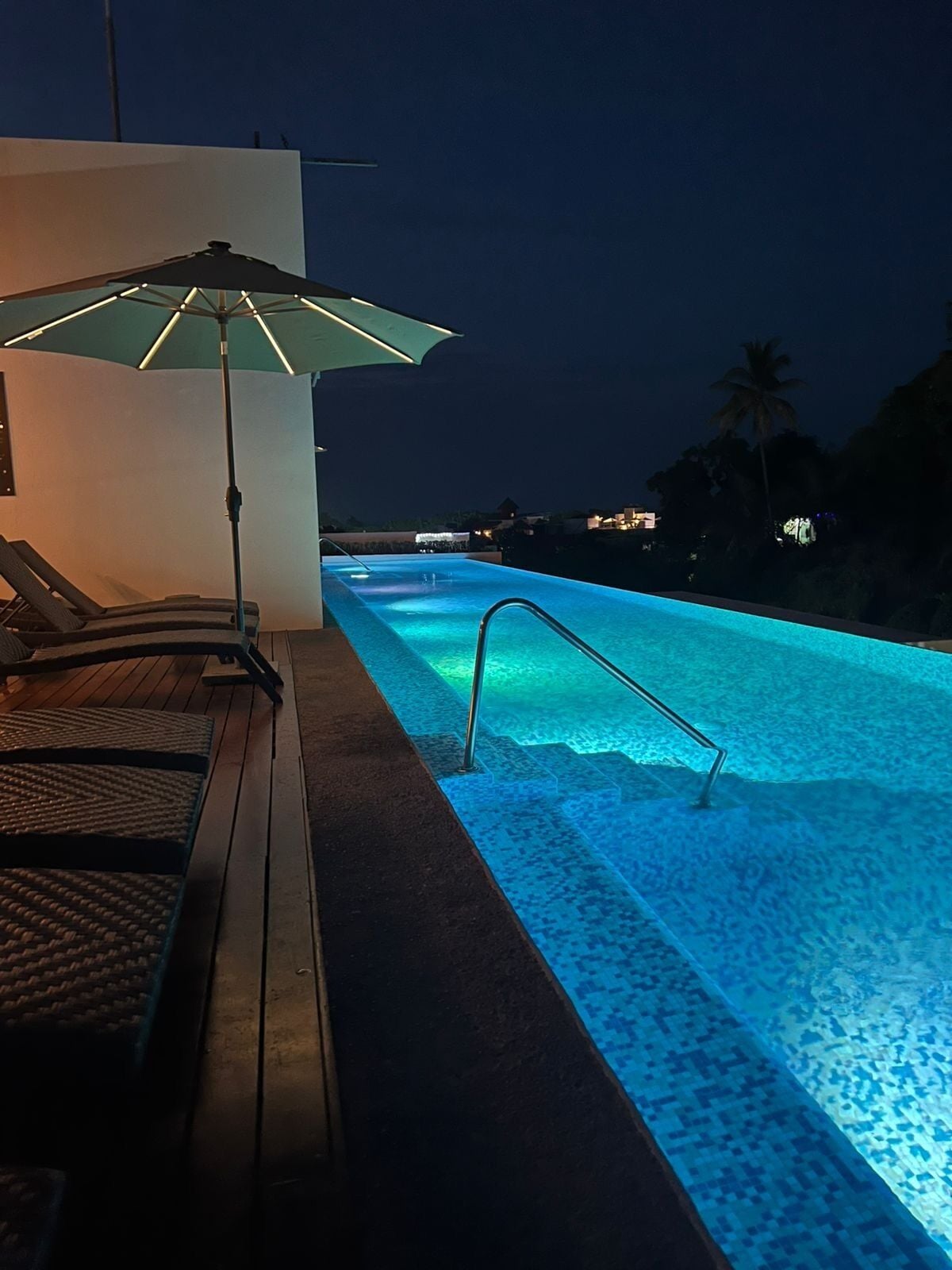 A nighttime swimming pool with a lounge area, a poolside umbrella, and a view of the surrounding area.