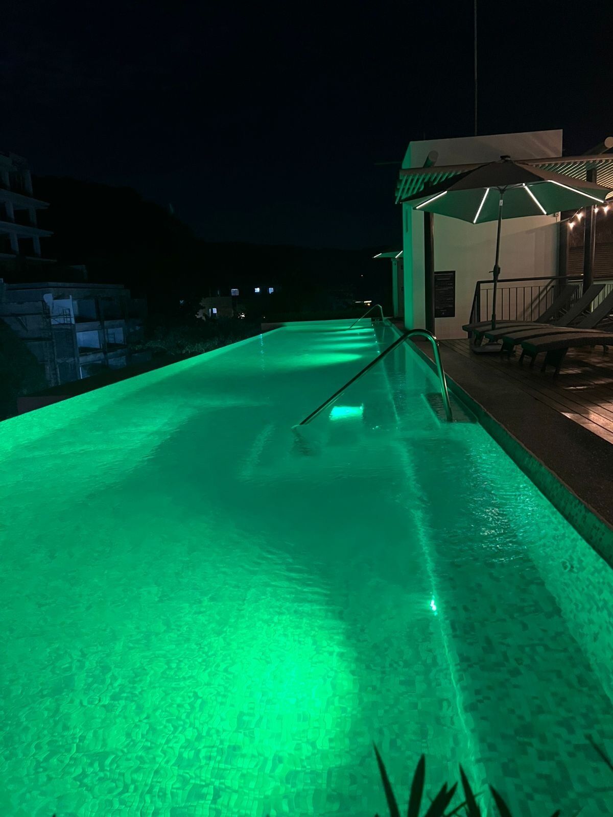 Una piscina con luz verde, un área de descanso con bancos y un edificio con porche cubierto.