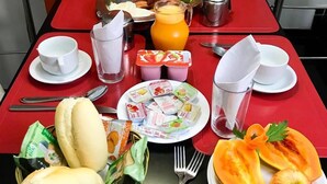 A breakfast setup with a red table, including a plate of food, a fruit bowl, a pitcher of orange juice, and various condiments. - Motel Villa di Roma - Adults Only (Guarujá)