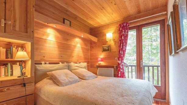 Une chambre à coucher dans un chalet en bois, avec un lit, une table de chevet, une lampe, une bibliothèque et une fenêtre avec des rideaux.