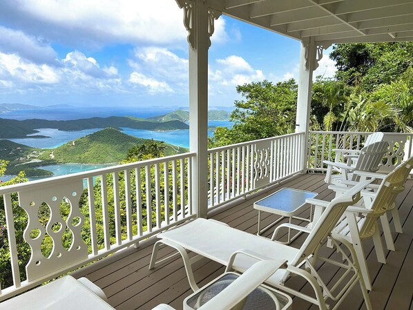 A covered outdoor seating area with white chairs and a table, overlooking a scenic landscape with hills and a body of water.
