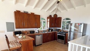 A kitchen with wooden cabinets, stainless steel appliances, and a dining area with a chair. - Experience the ultimate retreat at One Particular Harbour, where breathtaking views meet unparalleled privacy, creating the perfect oasis for relaxation and rejuvenation. This is the getaway you’ve been dreaming of! (Coral Bay)