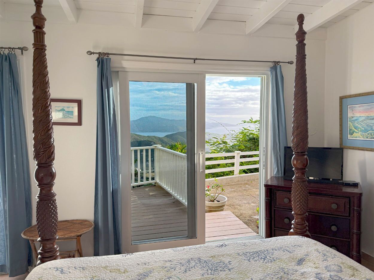 A bedroom with a four-poster bed, a wooden dresser, and a television. There is a balcony with a white railing and a view of the mountains.