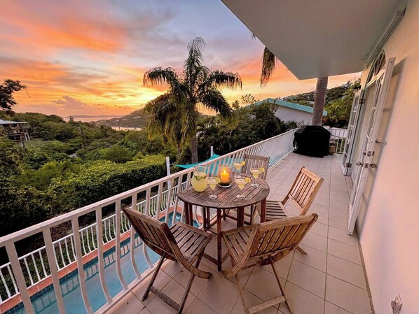 A balcony with a table and chairs, overlooking a swimming pool and lush greenery under a sunset sky.