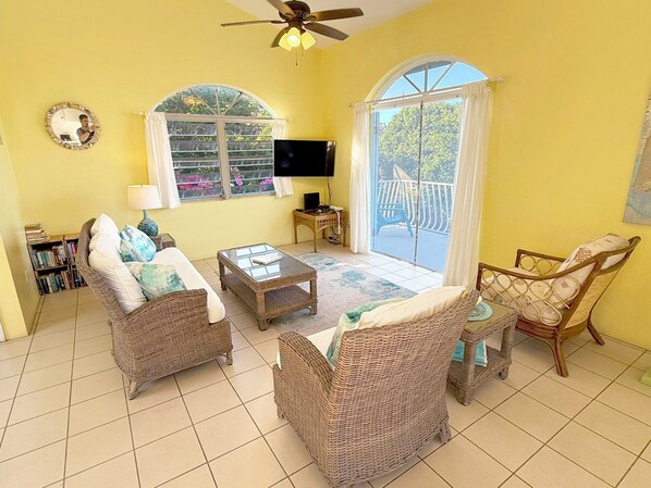 A room with wicker furniture, a ceiling fan, a flat-screen TV, and large windows with curtains.