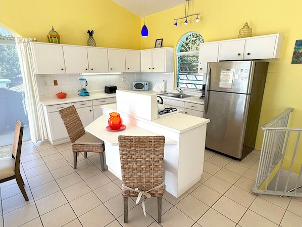 A compact kitchen with white cabinets, a stainless steel refrigerator, a microwave, and a small island with chairs.