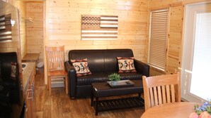 A cozy cabin interior with a wooden floor, a black leather sofa with decorative pillows, a wooden dining table, and a small plant on the coffee table.