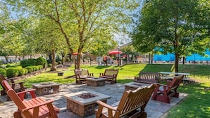 Un patio al aire libre con sillas Adirondack y mesas de piedra, rodeado de césped cuidado y árboles.
