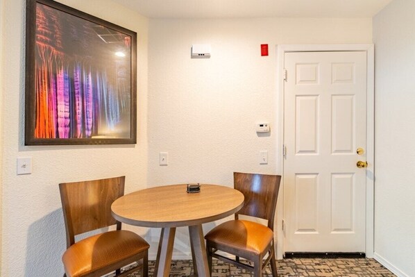 Una habitación con una mesa redonda de madera y dos sillas, un cuadro enmarcado en la pared, una puerta blanca y un equipo de aire acondicionado pequeño.