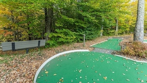 A golf course with a green artificial turf, a wooden bench, and a path surrounded by trees with autumn foliage.