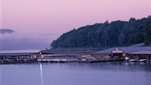 A waterfront property with a dock extending into calm water, a covered area with a red roof, and a forested hill in the background. - Wyndham Resort At Fairfield Bay - 1 Bedroom Deluxe (Fairfield Bay)