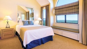 A hotel room with a large bed, two bedside lamps, a bench by the window, and a view of the sea. - Wyndham Resort At Fairfield Bay - 1 Bedroom Deluxe (Fairfield Bay)