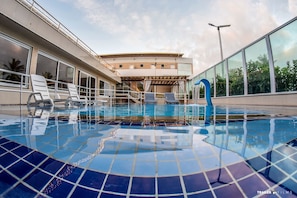Uma piscina com piso ladrilhado, cadeiras de lounge e um edifício com grandes janelas ao fundo.