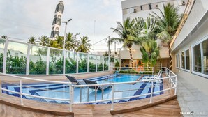 An indoor swimming pool with a tiled floor, surrounded by palm trees and a modern building with a clock tower in the background. - Farol Plaza Hotel (Aracaju)