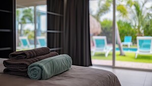Ein ordentlich bezogenes Bett mit gefalteten Handtüchern, ein großes Fenster mit Blick auf einen Pool und Liegestühle, und eine reetgedeckte Struktur im Hintergrund.
