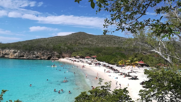 Ein Strand mit klarem türkisblauem Wasser, weißem Sand und einer bergigen Kulisse.